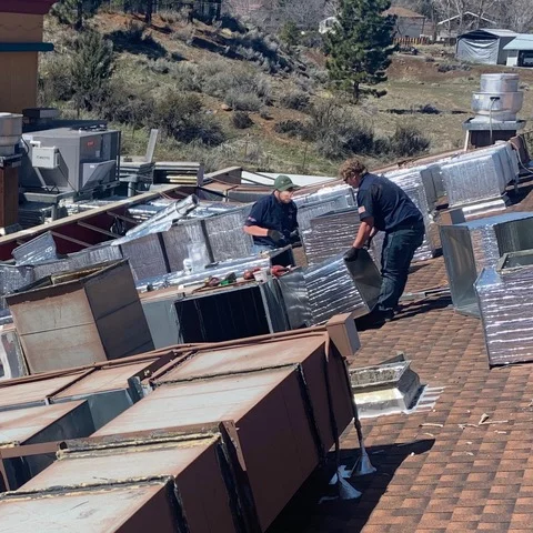 Two technicians installing metal HVAC units on asphalt shingle roof with multiple air conditioning systems