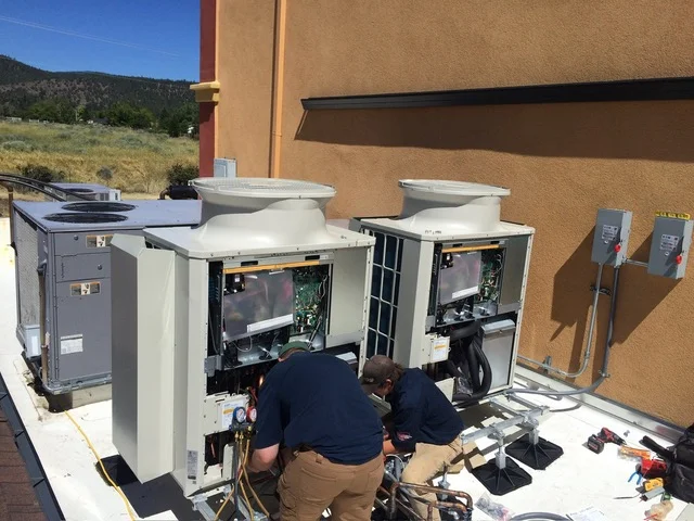 Two technicians installing dual mini-split heat pump outdoor units with exposed internal components and electrical connections